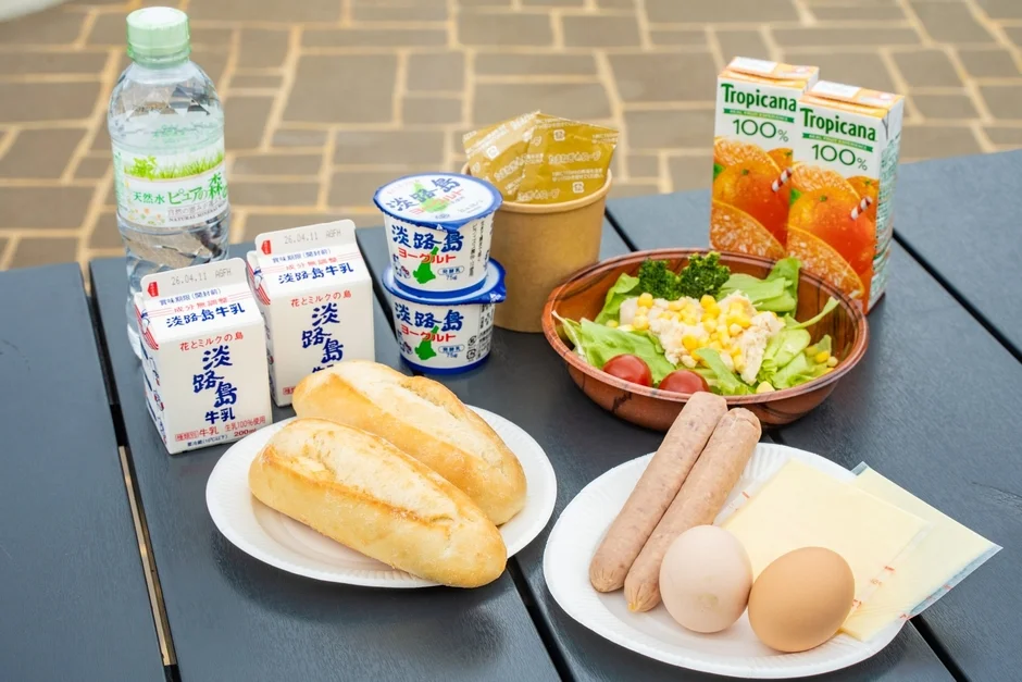 屋外のテーブルに並べられたバラエティ豊かな朝食セット。淡路島牛乳、ヨーグルト、トロピカーナジュース、天然水、サラダ、パン、ソーセージ、卵、チーズなどが写っています。