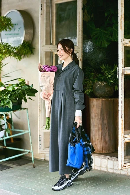 グレーのワンピースとスニーカー姿の女性が、紫色の花束と青いバッグを持ち、植物が置かれた店先で穏やかに微笑んでいます。