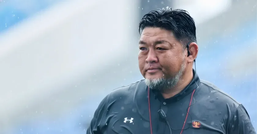 雨が降る屋外で、黒いスポーツウェアを着た中年男性が写っています。男性は真剣な表情で何かを見つめているようです。