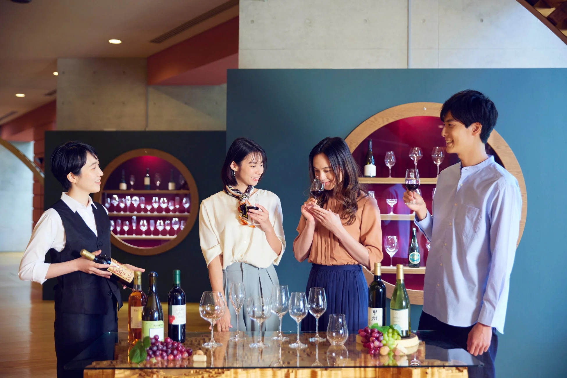 ワインショップまたは試飲会で、スタッフと3人の客がワインを楽しんでいる様子。テーブルには数種類のワインボトル、グラス、ブドウが並べられ、客はワインを試飲したり、説明を聞いたりしている。和やかな雰囲気でワイン文化に触れている様子が伺える。