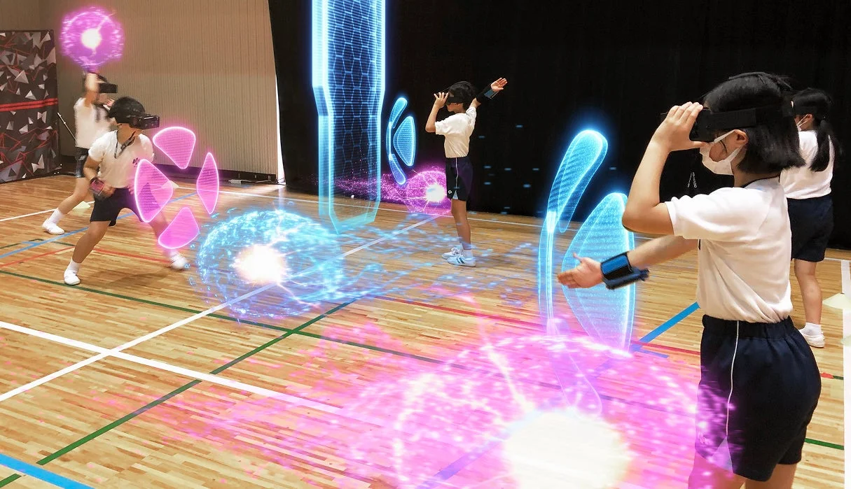 体育館で子供たちがVRヘッドセットと腕のデバイスを装着し、仮想空間の光るエフェクトとインタラクトしながら遊んでいる様子。未来的な教育やエンターテイメント体験を示唆する一枚です。