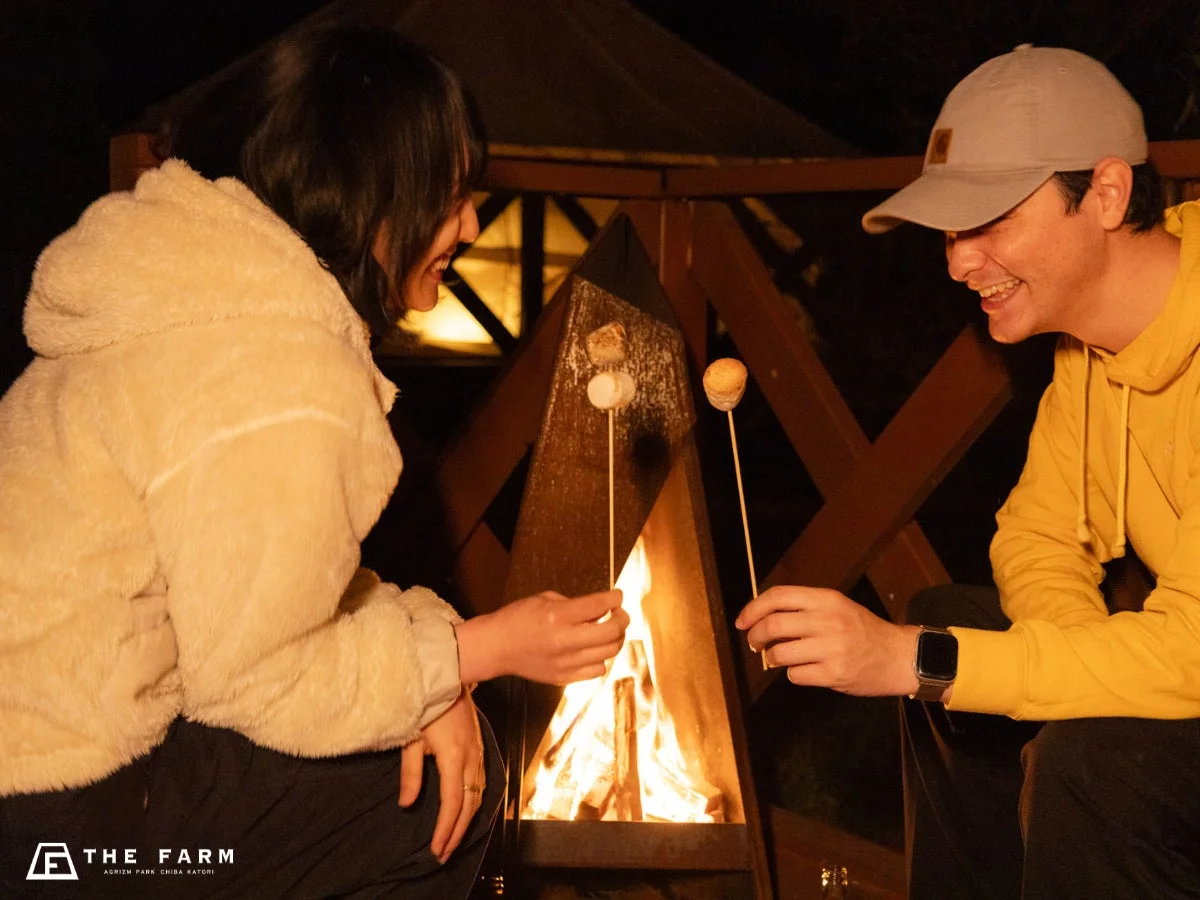 夜のキャンプ場で、男女のカップルが焚き火を囲んでマシュマロを焼いています。二人は笑顔で楽しそうに会話しており、暖かく和やかな雰囲気が伝わってきます。