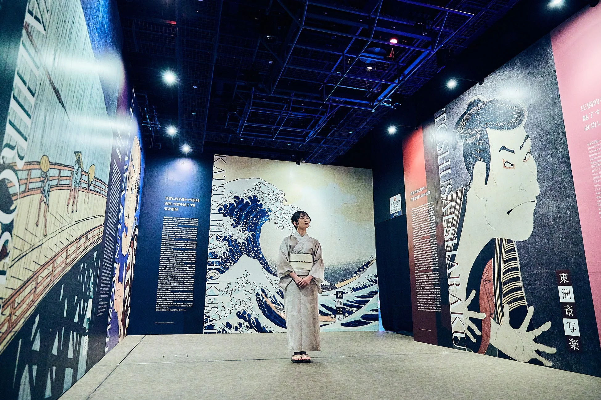 着物姿の女性が、葛飾北斎の「神奈川沖浪裏」や東洲斎写楽の役者絵などが大きく展示された空間に立っている。浮世絵をテーマにした現代的な美術展の様子がうかがえる。