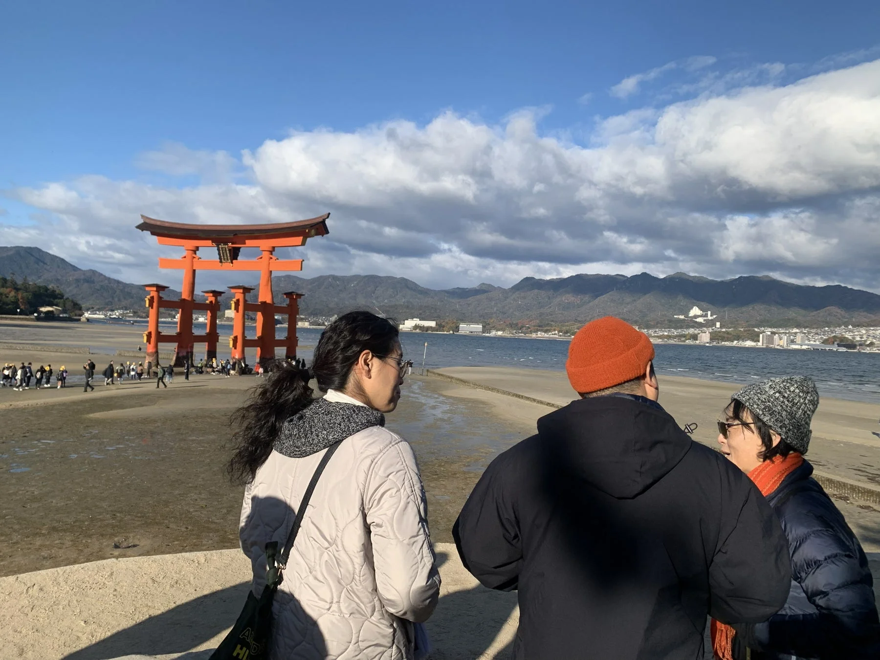 厳島神社の有名な大鳥居が干潮の砂浜に立つ姿を捉えた一枚です。晴れた空の下、数人の観光客が大鳥居や海の景色を眺めています。背景には穏やかな海と連なる山々が広がり、日本の美しい風景が感じられます。