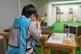 屋内射撃場で、一人の人物がライフルを構え、標的を狙って射撃練習をしている様子です。