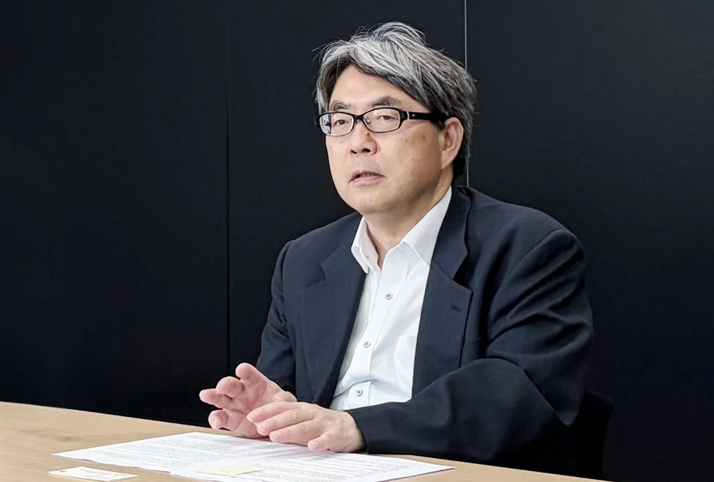 眼鏡をかけた中高年の男性が、白いシャツとスーツを着てテーブルに座っています。何かを説明しているかのように手ぶりをしており、ビジネスシーンでの会議やインタビューの様子を捉えた写真です。