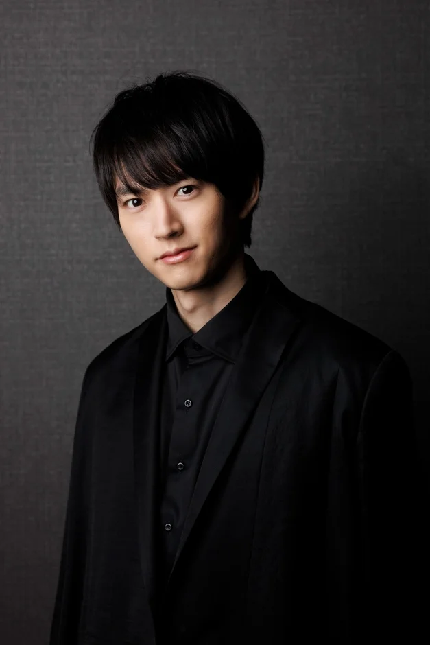 黒い髪の若い男性が黒いシャツとジャケットを着用し、カメラに向かって穏やかに微笑んでいます。