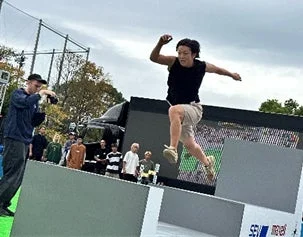 屋外のイベント会場で、黒いタンクトップ姿の男性が白い障害物を飛び越えている瞬間を捉えた画像です。