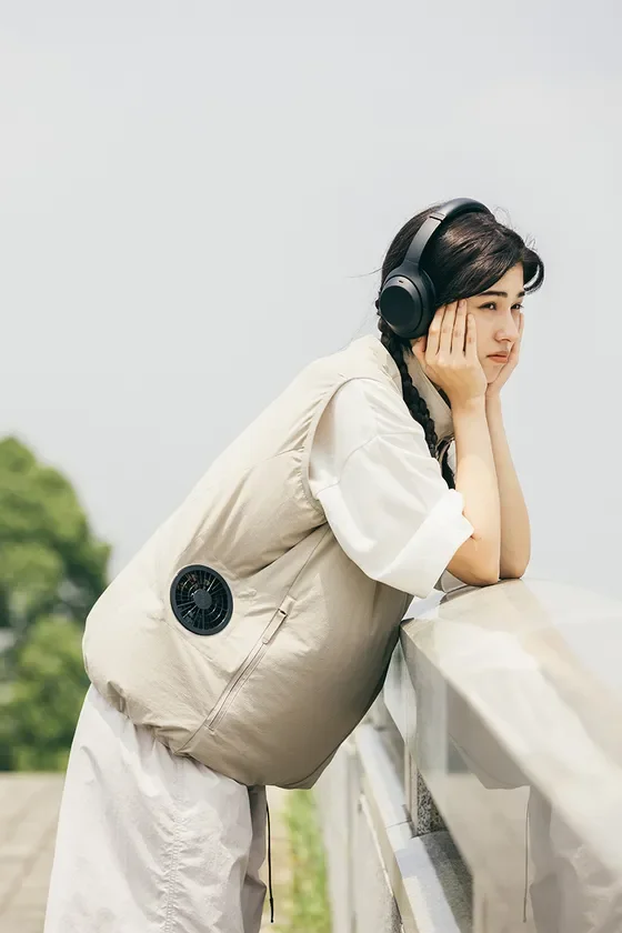 明るい屋外で、ヘッドホンを装着し、ファン付きベストを着た若い女性が手すりに寄りかかっている。遠くを見つめ、物思いにふけるような表情をしている。涼しげな夏のファッションスタイルが特徴的。