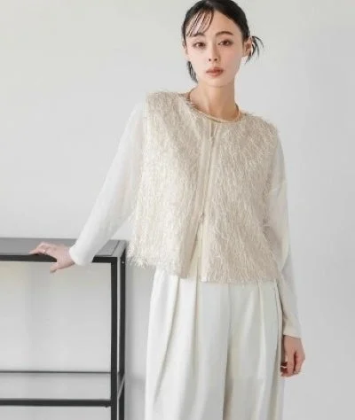 白い長袖トップスにベージュのフリンジベストと白いワイドパンツを合わせた女性の全身ポートレート。ミニマリストな背景で、洗練されたファッションスタイルを表現しています。