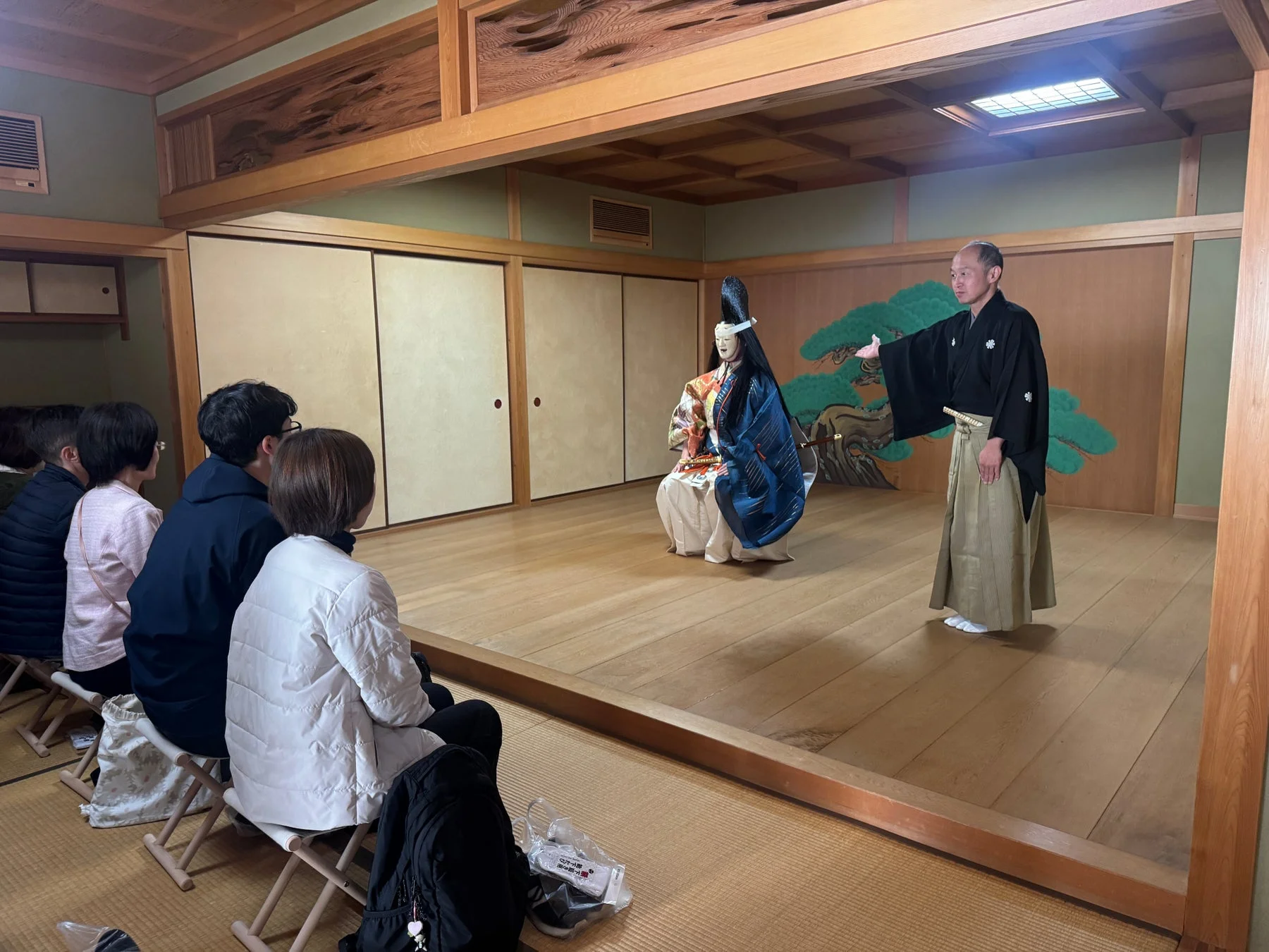 和室で日本の伝統芸能が披露されており、観客が熱心に見入っています。和服を着た人物が大きな人形を指し示し、舞台の背景には松の絵が描かれています。