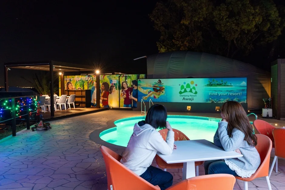 夜のリゾート施設で、ライトアップされたプールサイドに2人の女性が座っています。背景には「Glamping Resort Awaji」の広告看板があり、リラックスした休暇の雰囲気が漂っています。