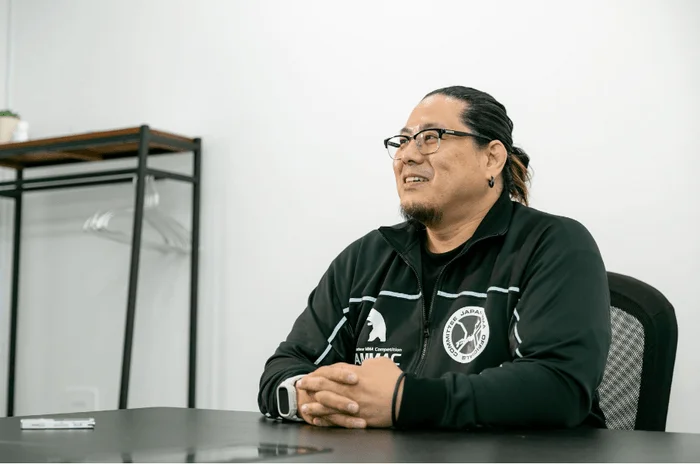 メガネをかけた男性が、室内でテーブルに座り笑顔を見せています。黒いジャケットには「COMMITTEE JAPAN OFFICIALS」や「MMA Competition AMMAC」の文字が見えます。会議中か、インタビューを受けているような様子です。