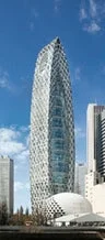 青空の下にそびえ立つ、独特な幾何学模様の外観を持つ高層ビルディングです。周囲には他の都市の建物も見えます。