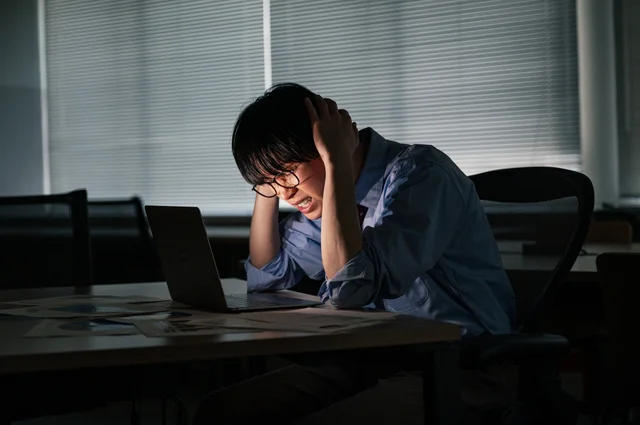 夜のオフィスで、パソコンに向かい頭を抱え、困惑した表情を浮かべる男性