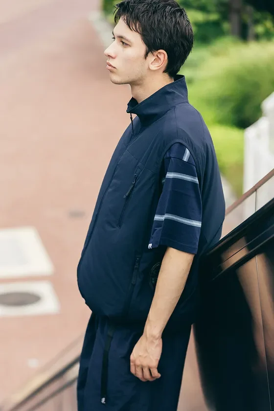 ネイビーのベストとストライプの半袖Tシャツを着用した若い男性が屋外で横向きに立っている写真です。カジュアルでスポーティなファッションが特徴的で、背景にはエスカレーターのような構造が見えます。