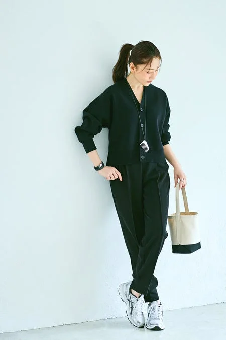 黒のVネックカーディガンとパンツを合わせたモノトーンコーデの女性が、白いスニーカーとトートバッグを持って立っている。