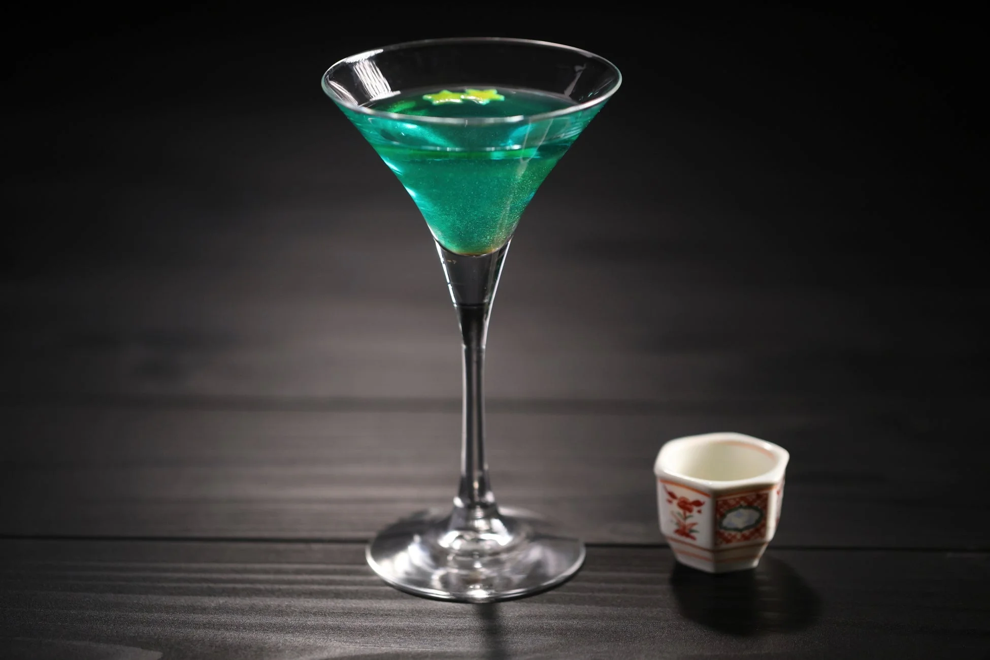 きらめく青緑色のカクテルと和風の小さな杯