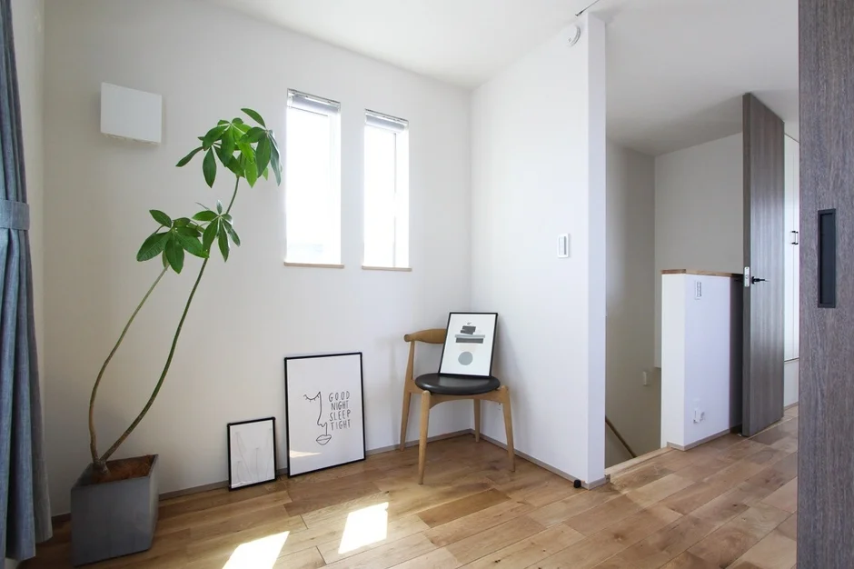 白い壁と明るいフローリングが特徴のシンプルモダンな空間です。観葉植物、木製の椅子、アートが配され、奥には階段とドアが見えます。自然光が差し込む清潔感あふれるインテリアです。