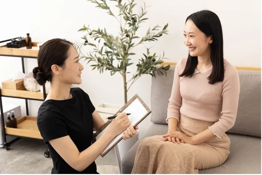 明るい室内で二人のアジア人女性が向かい合って話しています。一人はクリップボードに何かを書き込みながら、もう一人の女性と笑顔で会話しており、カウンセリングやコンサルティングの場面のように見えます。