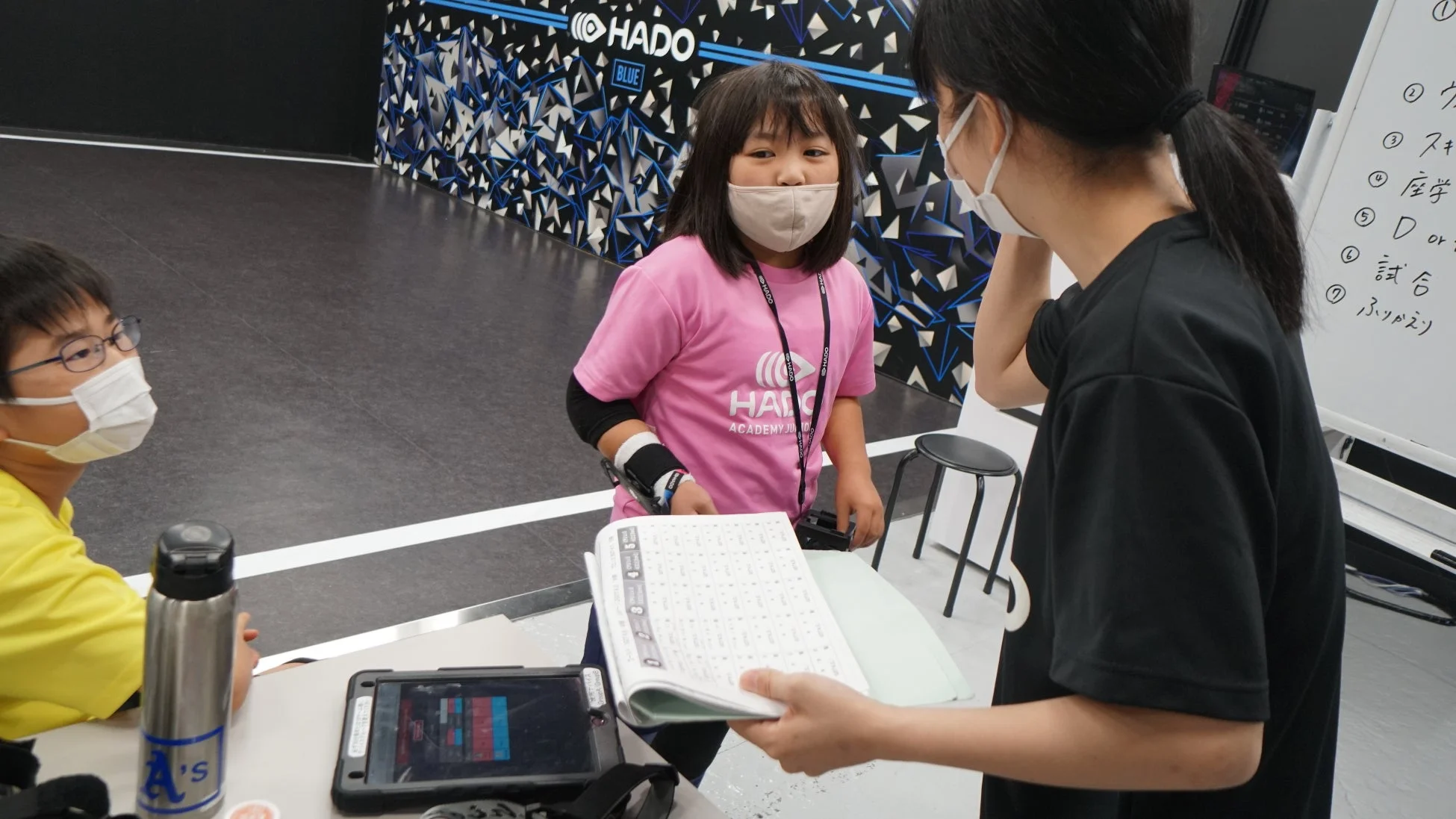 HADOイベントでマスクを着用した子供たちと大人が交流している様子。HADOアカデミーのTシャツを着た少女が、資料を見ながら説明を受けている。奥にはHADOのロゴとホワイトボードが見える。