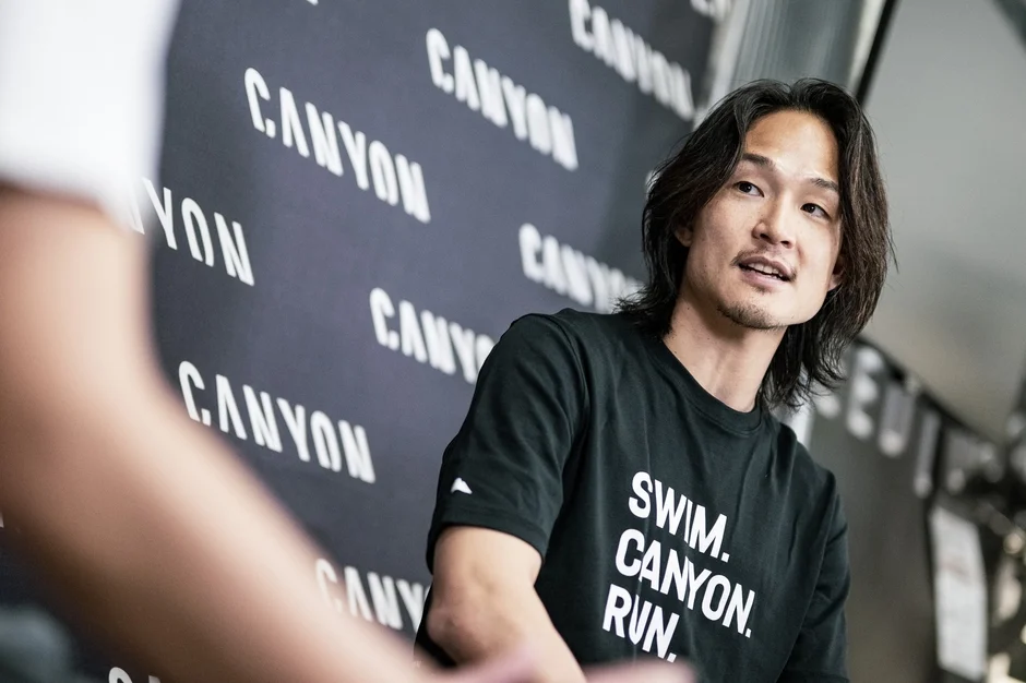 「CANYON」のロゴが並ぶ壁を背景に、黒いTシャツを着たアジア人男性が話している。Tシャツには「SWIM. CANYON. RUN.」の文字があり、スポーツ関連のイベントやインタビューの様子を捉えた一枚。