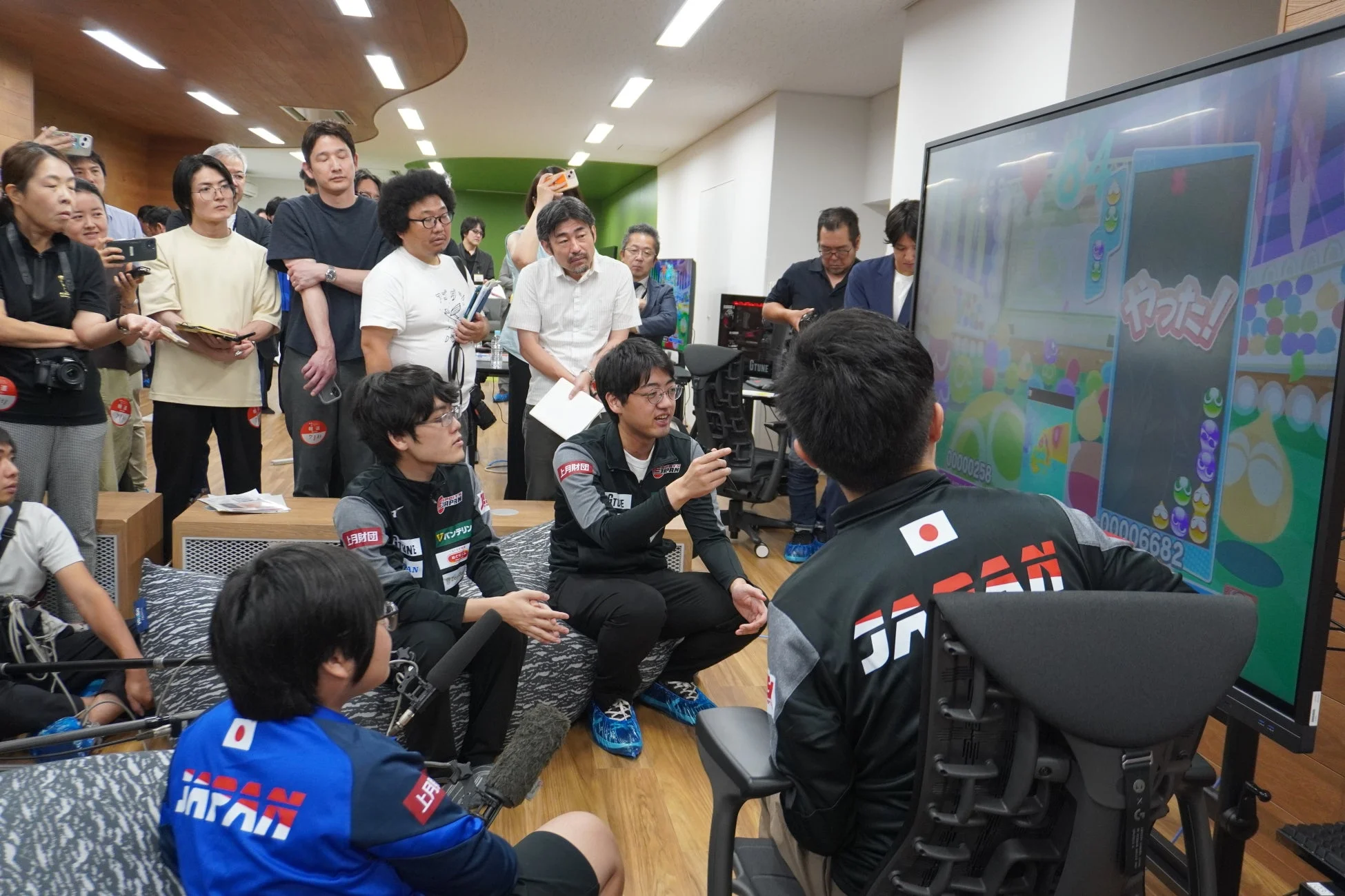 「JAPAN」のロゴが入ったウェアを着た若者たちが、大型モニターに映し出されたゲーム画面を見ながら真剣に議論している様子