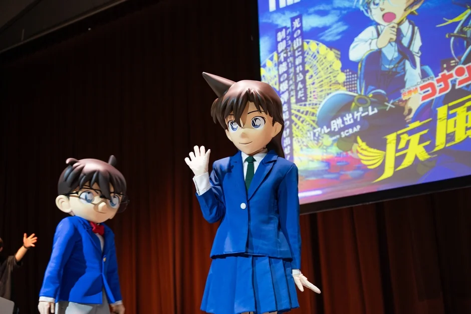 名探偵コナンの江戸川コナンと毛利蘭の着ぐるみキャラクターがステージに立っており、背景のスクリーンには「リアル脱出ゲーム」の告知が映し出されています。