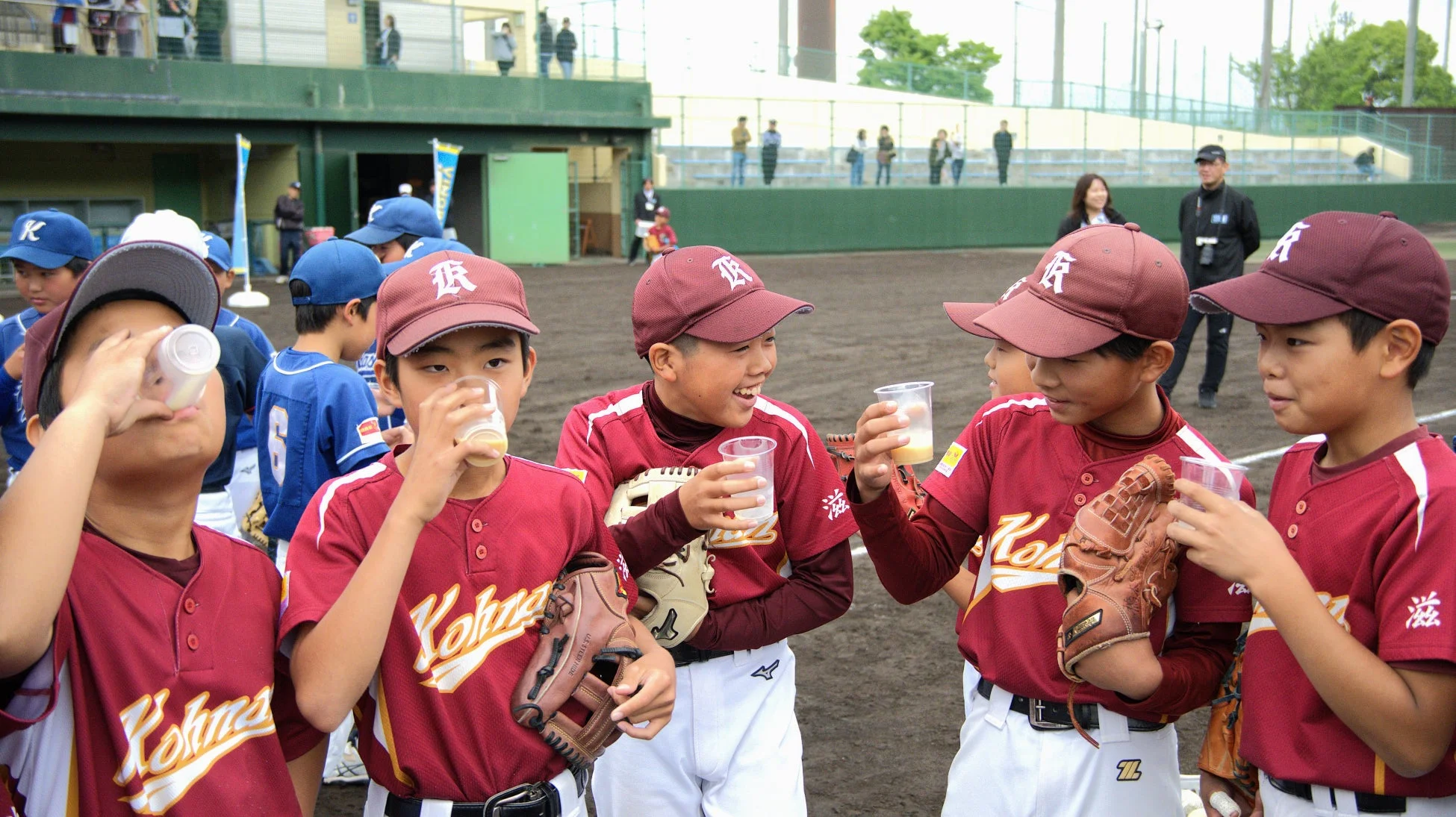 野球のユニフォームを着た少年たちが、グラウンドで休憩中に飲み物を飲んでいる様子。数人の子供たちが笑顔を見せており、スポーツの楽しさやチームメイトとの絆が感じられます。