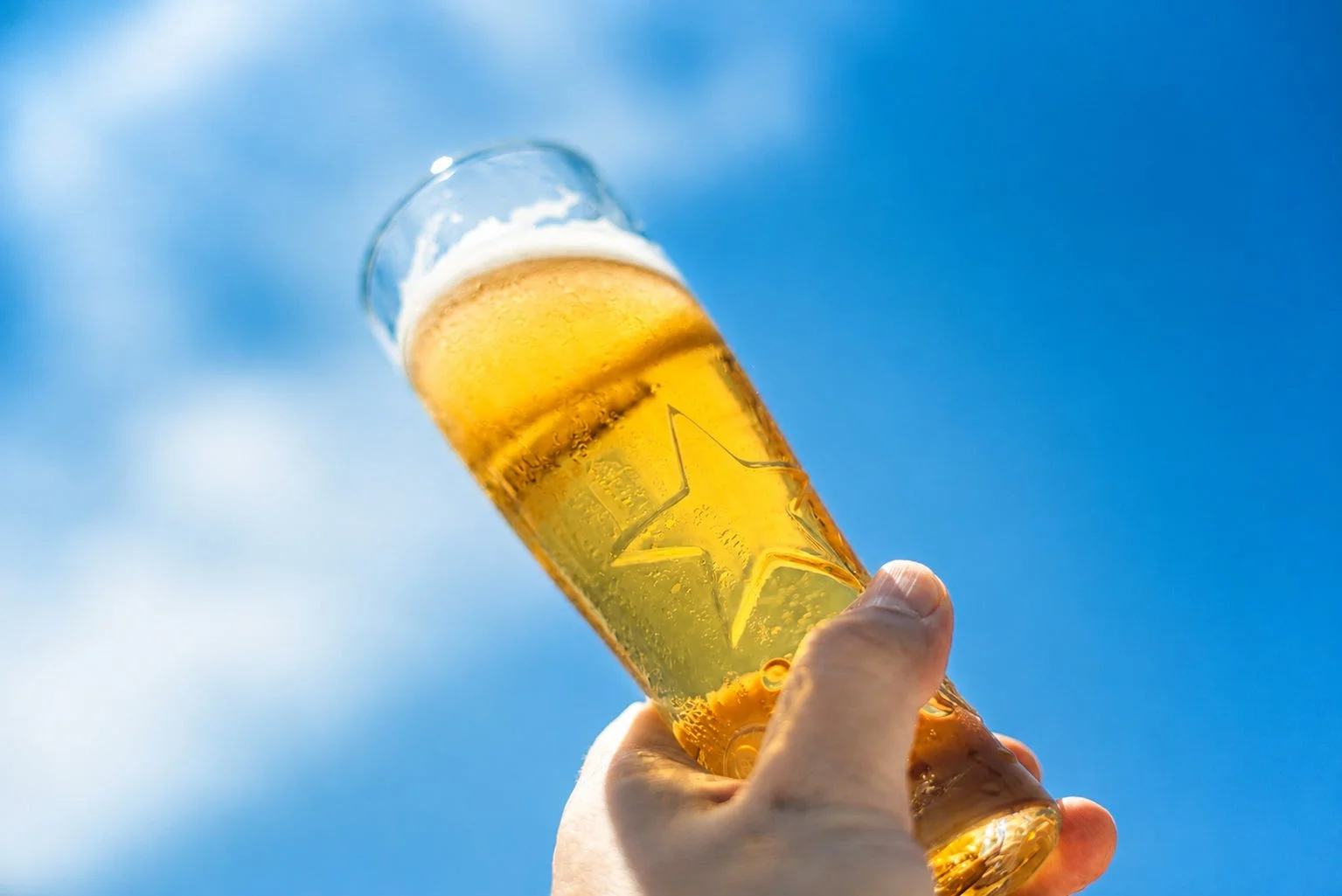 晴れた青空を背景に、泡が乗った冷たいビールグラスを掲げている手のクローズアップ。夏の日差しの中で飲むビールの爽快感を表現しています。