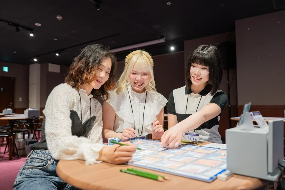 3人の若い女性がテーブルを囲み、笑顔で何かの活動に取り組んでいます。