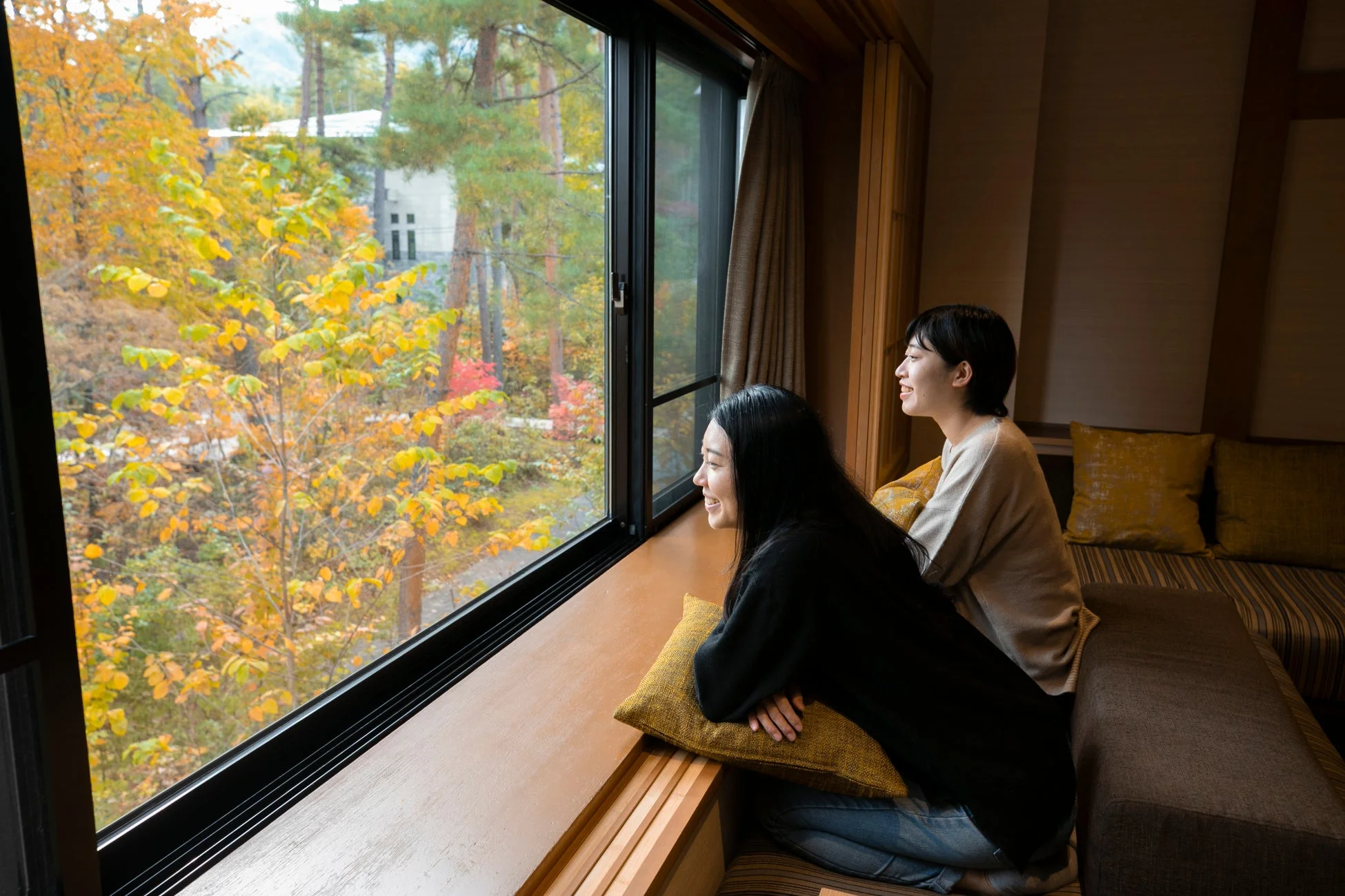 窓辺に座る二人の女性が、紅葉に彩られた美しい秋の景色を眺め、笑顔でリラックスしている様子。ホテルの一室のような落ち着いた空間で、自然を満喫している。