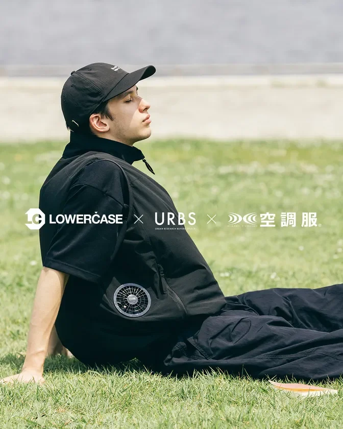 男性が黒い空調服とキャップを着用し、屋外の芝生に座ってリラックスしている様子。LOWERCASE、URBS、空調服のコラボレーションアイテムを着用しており、夏の快適なファッションを提案。