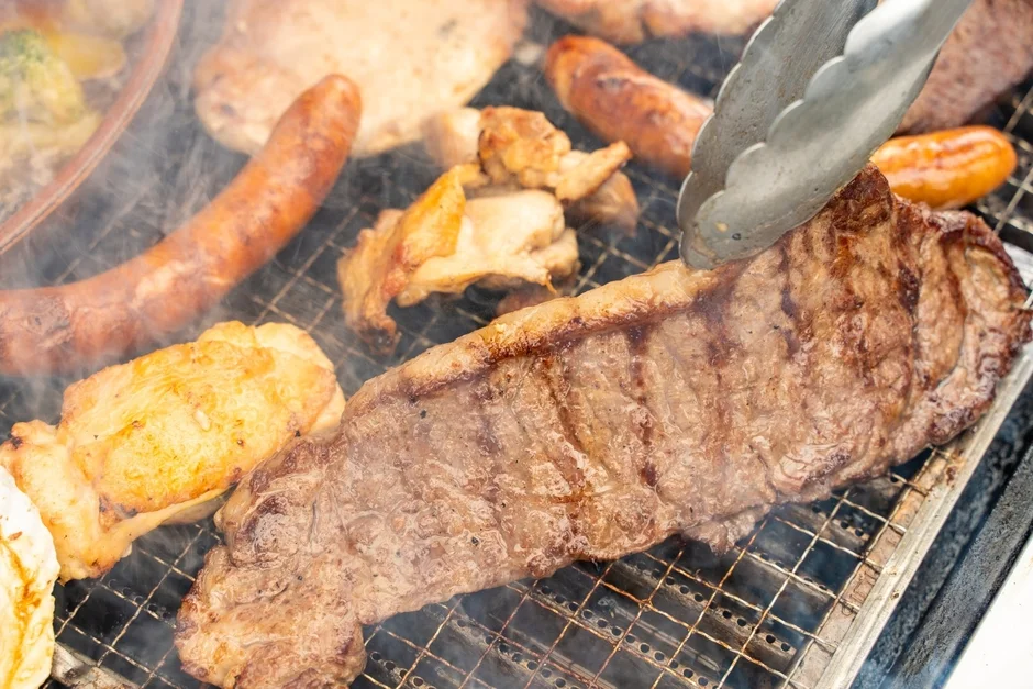 熱い網の上でステーキやソーセージ、鶏肉などが焼かれており、煙が立ち上るバーベキューの様子を捉えた写真です。トングで肉をひっくり返している瞬間が写っています。