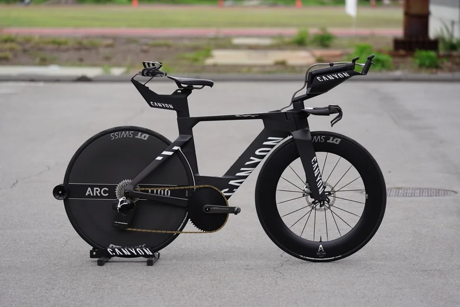 黒いCANYON製タイムトライアルバイクが舗装路に停車しています。空気抵抗を最小限に抑えた流線形のフレームデザインと、後輪のディスクホイール、前輪のディープリムホイールが特徴で、高性能な競技用自転車です。