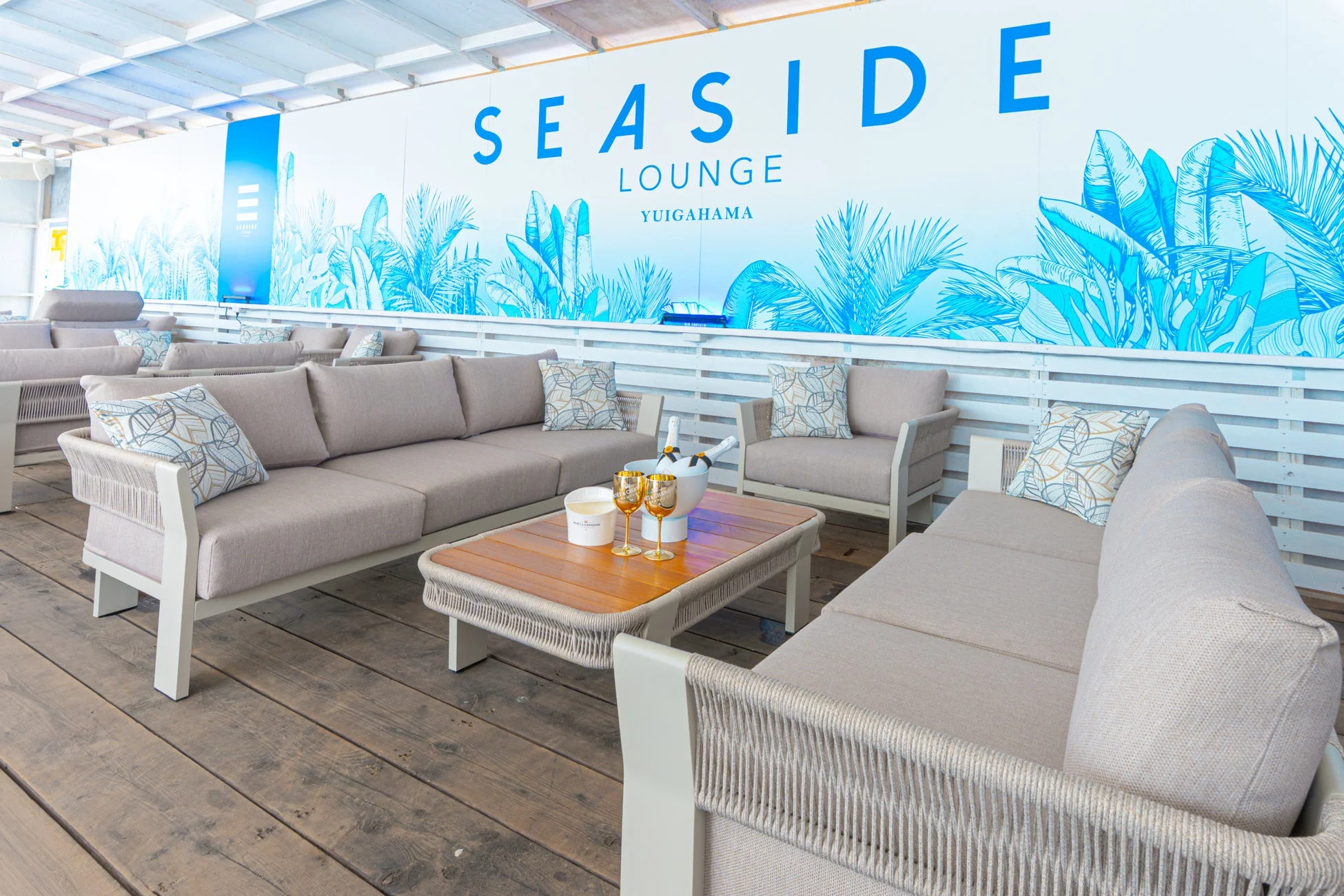 「SEASIDE LOUNGE YUIGAHAMA」と書かれた壁が特徴的な、屋外のスタイリッシュなラウンジエリアです。快適なソファとシャンパンが置かれたテーブルがあり、リゾートのような雰囲気でくつろげます。