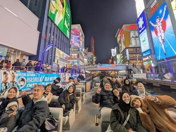 夜の道頓堀で、多くの観光客が運河のボートツアーを楽しんでいる様子です。グリコの看板やアサヒビールの広告など、華やかなネオンが街を彩り、活気あふれる日本の夜の風景が広がっています。