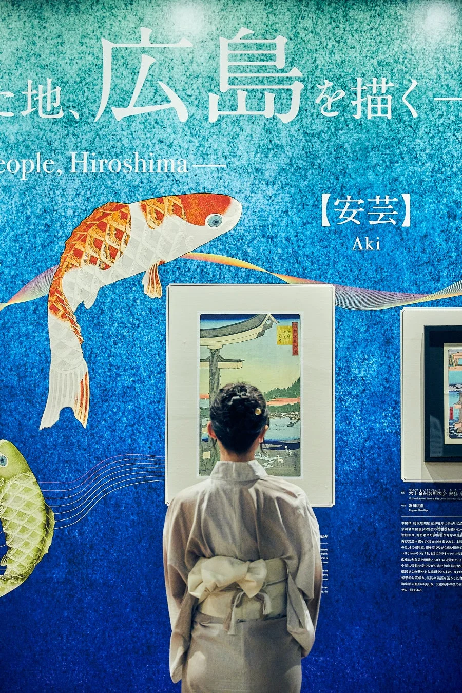 着物を着た人物が、青い背景に鯉のイラストが描かれた展示壁の前で、歌川広重の浮世絵を鑑賞している様子。広島をテーマにした展覧会の風景。