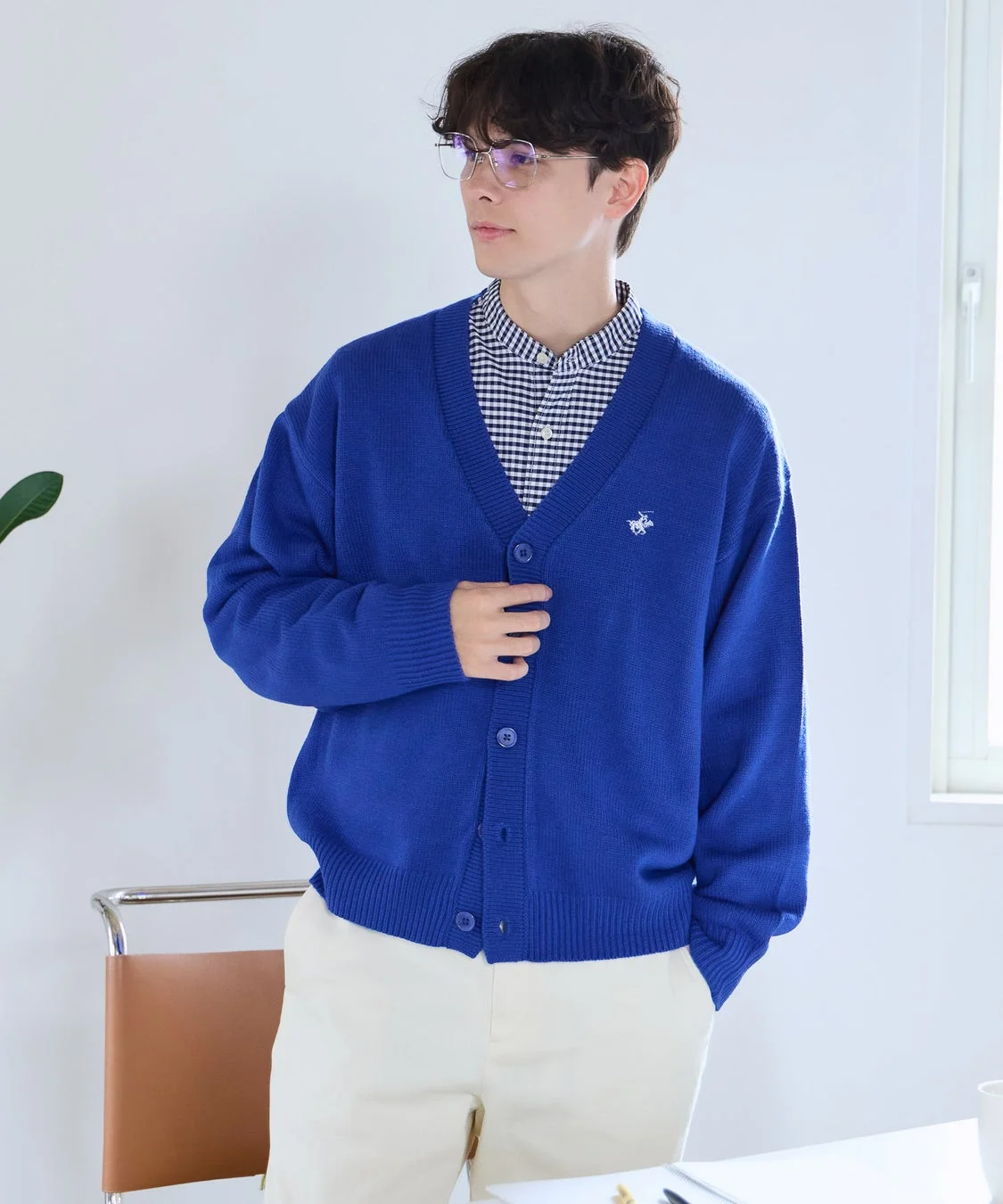 青いVネックカーディガンとチェックシャツを着用した男性が、メガネをかけ、屋内でポーズを取っている