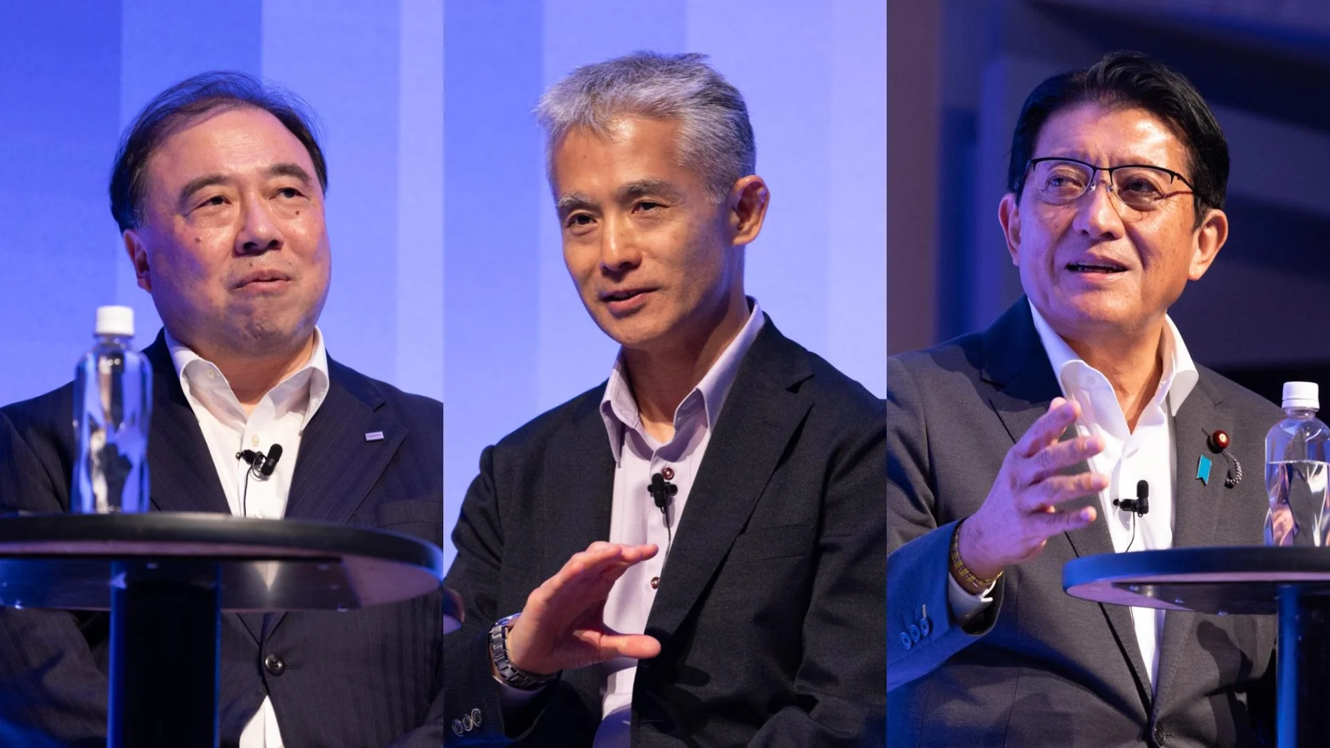 3人の男性が壇上で会議またはパネルディスカッションに参加している様子。それぞれスーツを着用し、マイクと水ボトルが置かれたテーブルに着席しており、真剣な表情や話している様子がうかがえます。ビジネスイベントの一場面と見られます。