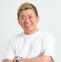 白いTシャツを着て腕組みをし、笑顔でカメラを見つめる金髪の中年男性のポートレート