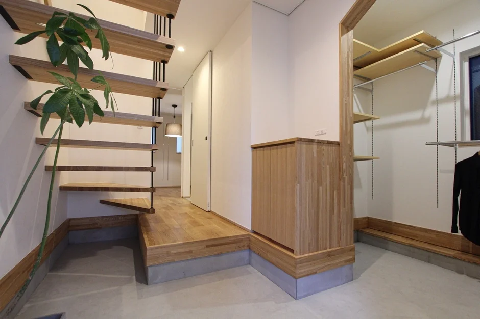 モダンな住宅の明るい空間。木製のオープン階段と観葉植物が特徴的で、奥には木目調の収納とオープンクローゼットが見える。機能的で洗練されたデザインの内装。