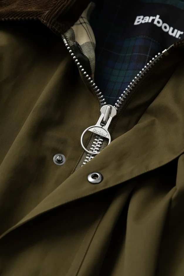 オリーブ色のBarbourジャケットのアップで、ジッパーの引き手と内側にブランドロゴが見えます。襟は茶色のコーデュロイ、裏地はチェック柄で、細部のデザインと素材感がよく分かる写真です。