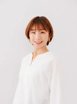白い背景の前で、笑顔でカメラを見つめるショートヘアの女性のポートレート。白いVネックのシャツを着用し、シンプルなアクセサリーを身につけている。明るく親しみやすい印象。