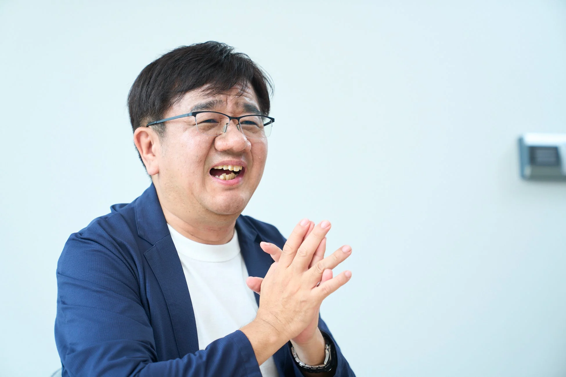 メガネをかけた中年の日本人男性が、明るい笑顔で手を組みながら話している様子