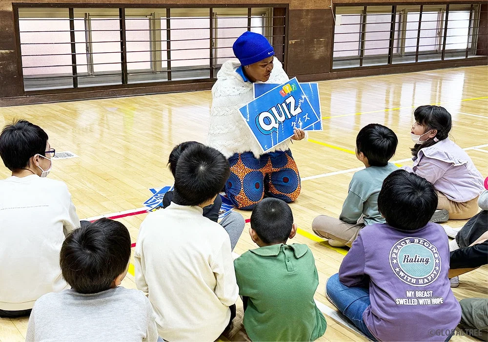 体育館で、一人の大人が子供たちを前にクイズボードを持って説明しています