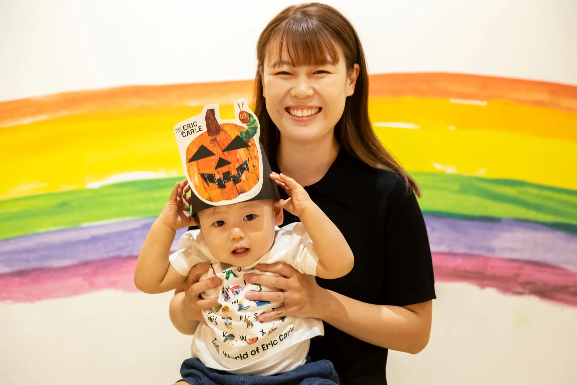笑顔の女性が、エリック・カール作のハロウィンかぼちゃの帽子をかぶった赤ちゃんを抱いています。