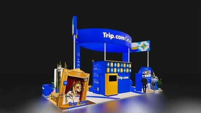 Trip.com Groupの青と黄色のモダンな展示ブースが写っています。ブースには「CIAO VENEZIA」と書かれた金色の額縁の中にベネチア風の仮面が飾られており、旅行関連のプロモーションイベントの様子を伺わせます。