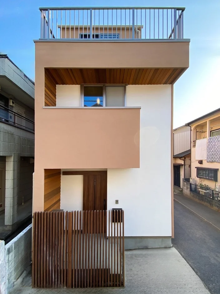 青空の下に建つ、白とベージュのモダンな外観が特徴的な狭小住宅です。屋上には手すり付きのバルコニーがあり、玄関前には木製の縦格子フェンスが設置されています。