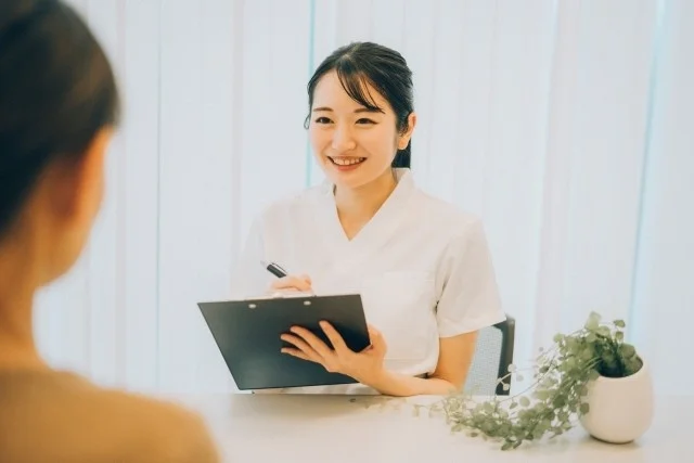 白い制服を着た女性が、笑顔でクリップボードにペンで書き込みながら、目の前の人物と対話している様子